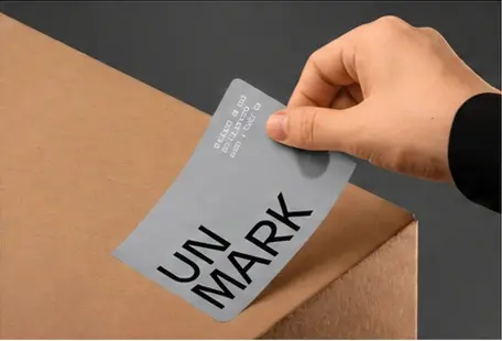 A hand placing a gray UN MARK brand label onto a cardboard box.